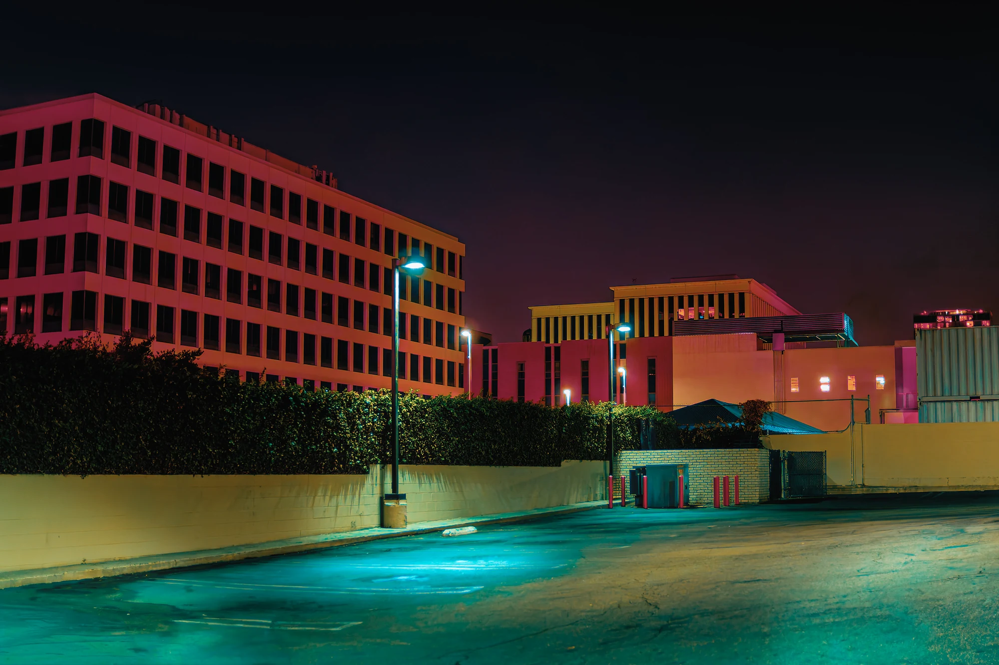 Pink-lit office buildings behind a hedge and low wall at night, an empty parking lot glowing cyan in the foreground.