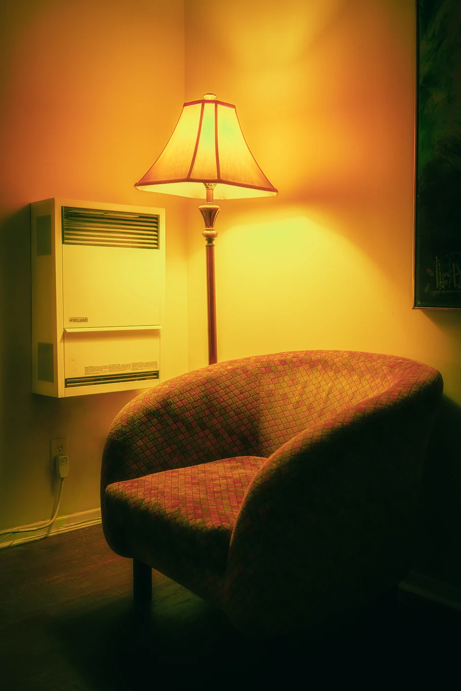 Fauteuil à motifs vide dans l’angle d’une chambre éclairée par un lampadaire, un radiateur mural blanc à côté, le tout dans une lumière ambrée.