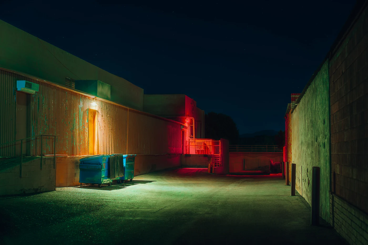 Empty service alley behind buildings at night, blue dumpsters under green light and a staircase glowing red at the far end.