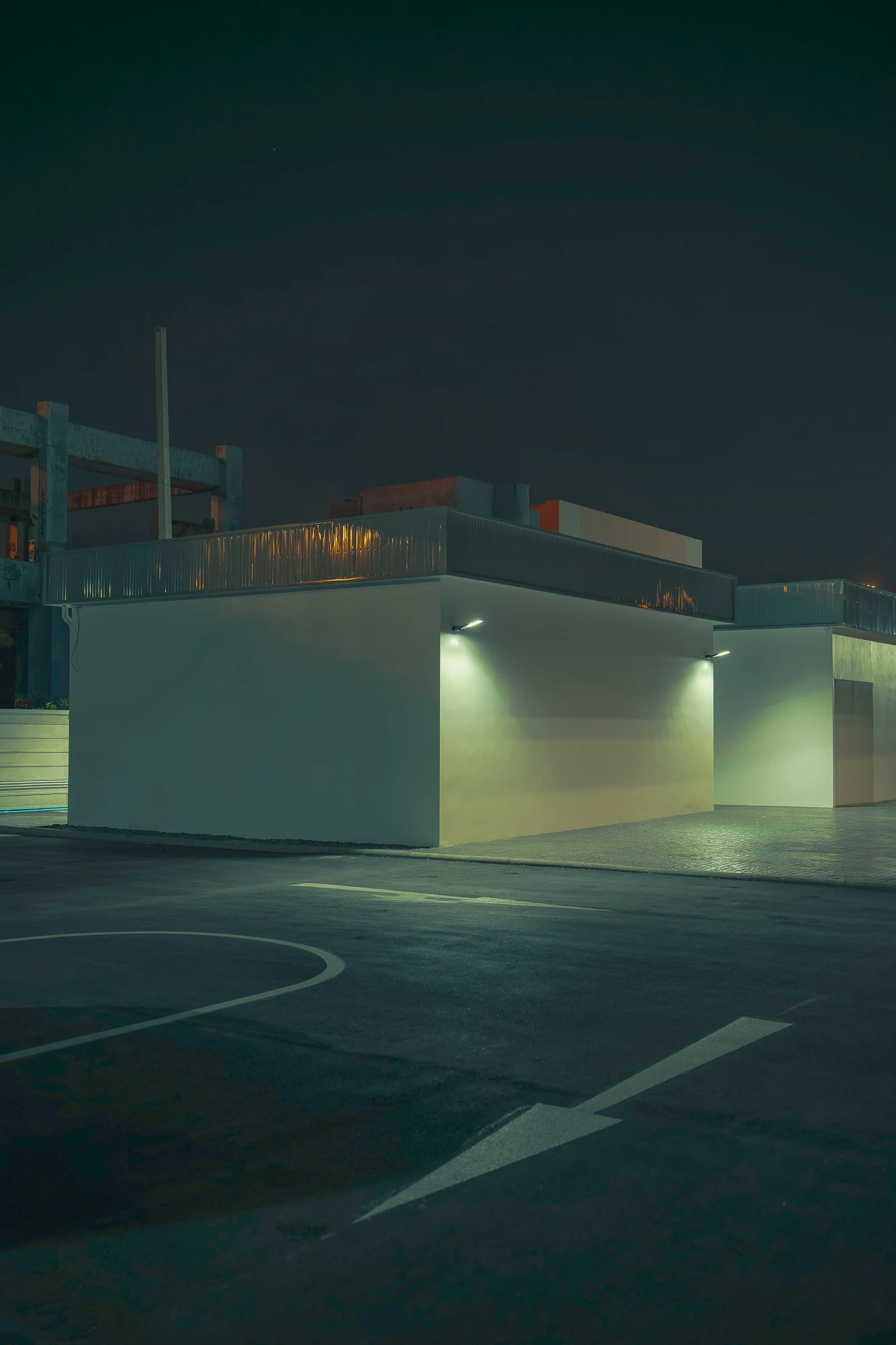 White windowless buildings lit by small wall lamps beside an empty asphalt court with painted lines and an arrow at night.