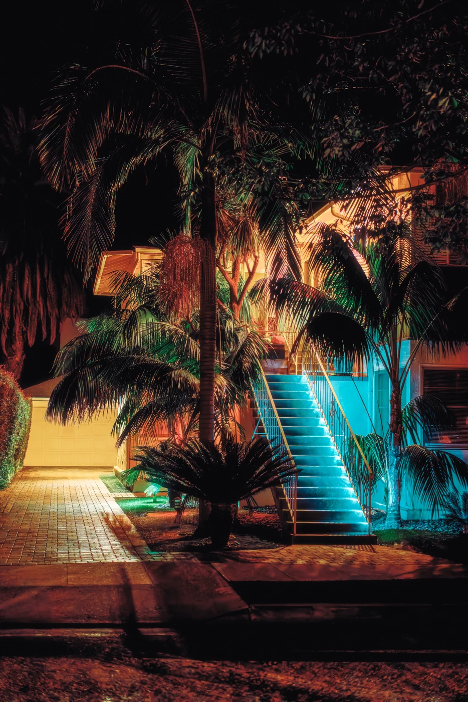 Exterior staircase glowing cyan among palm trees in front of a house at night, warm light on fronds and a paved driveway.
