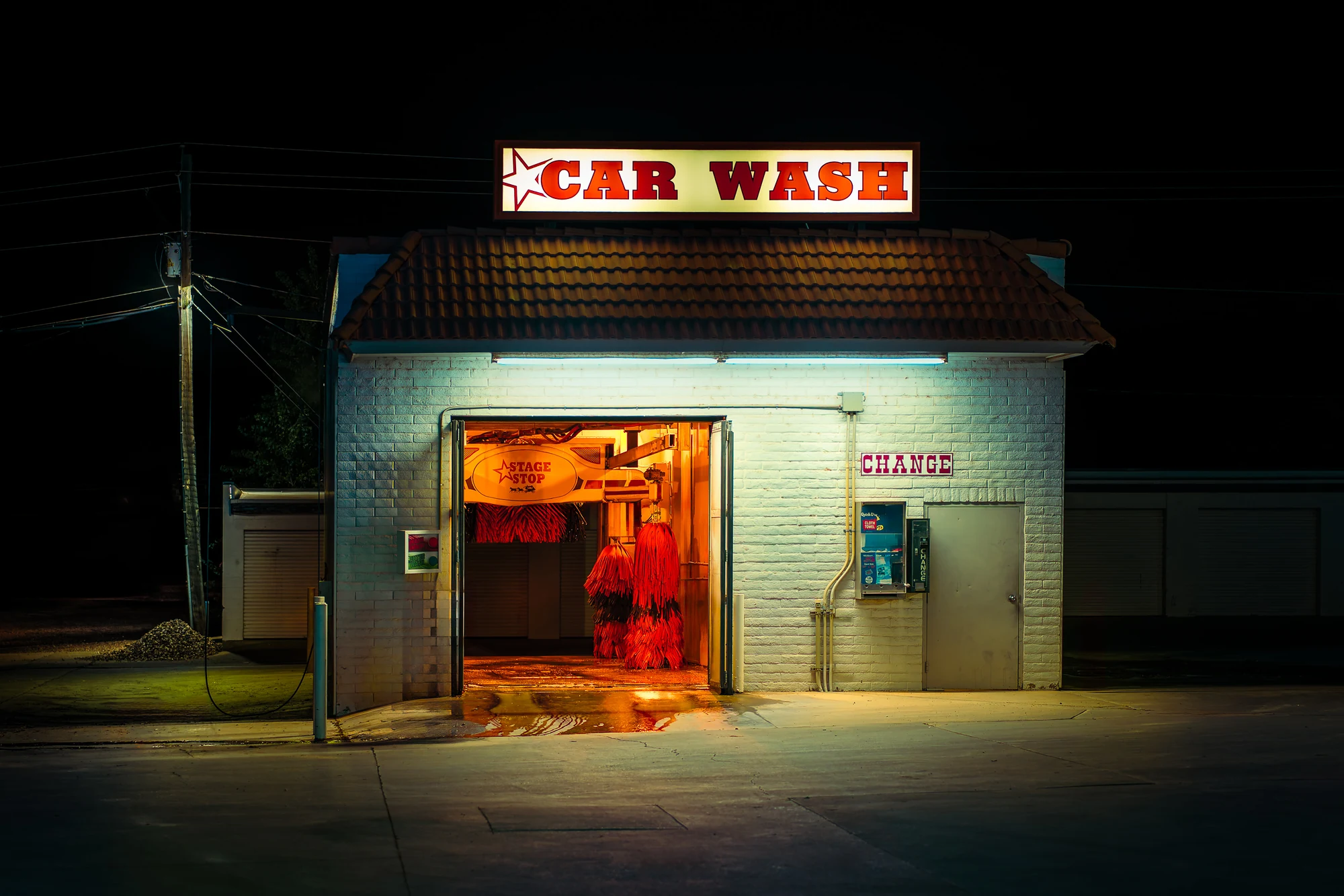 Small car wash lit at night, red "CAR WASH" sign on the roof, open bay showing red washing brushes in warm orange light.