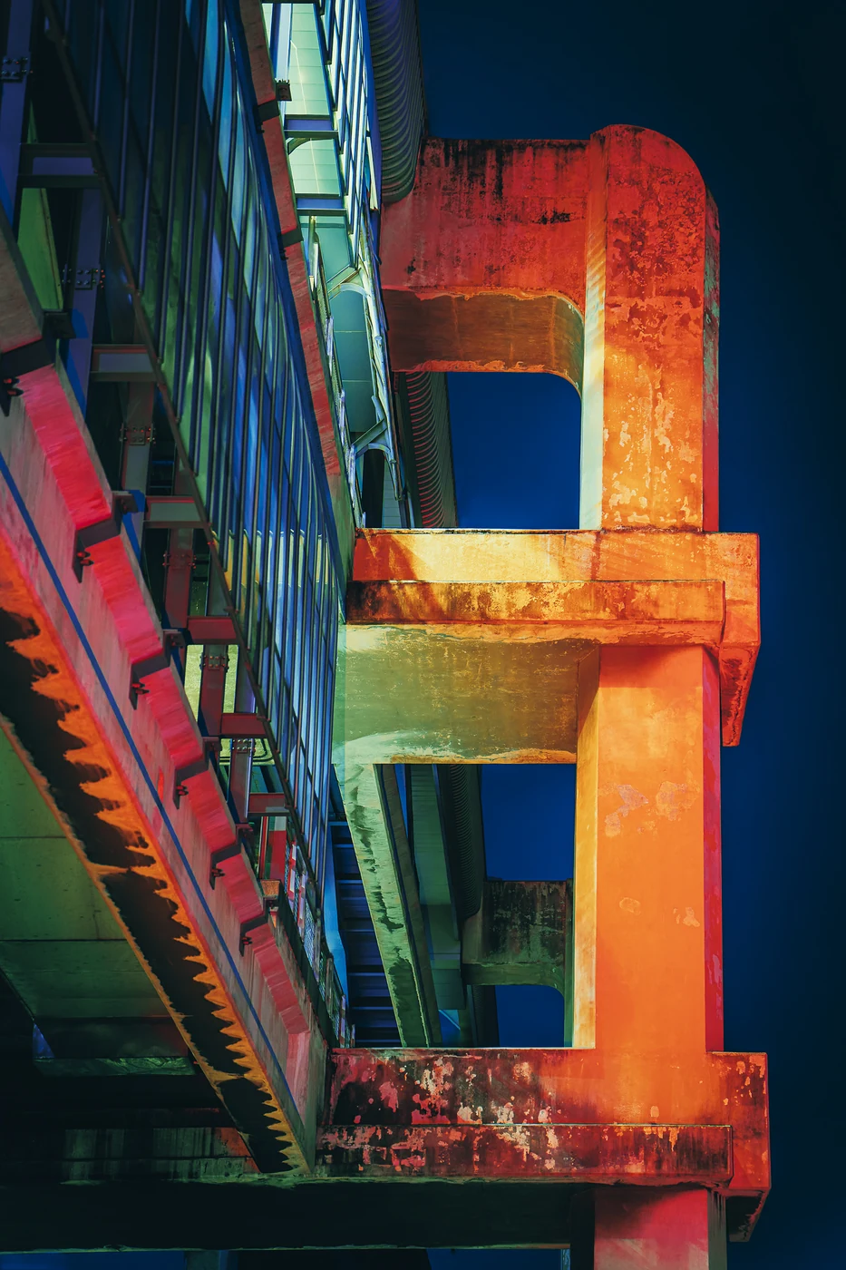 Piliers de béton usés d’une structure surélevée éclairés en orange et rouge sur un ciel bleu profond, une passerelle vitrée à côté.