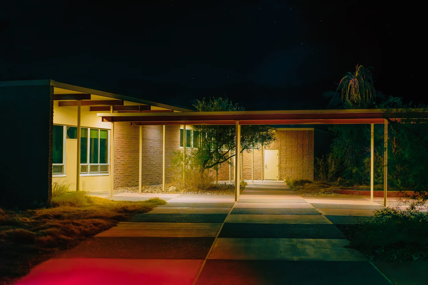 Bâtiment bas des années 1950 avec une longue galerie couverte éclairée la nuit, une lueur rouge sur le sol au premier plan.