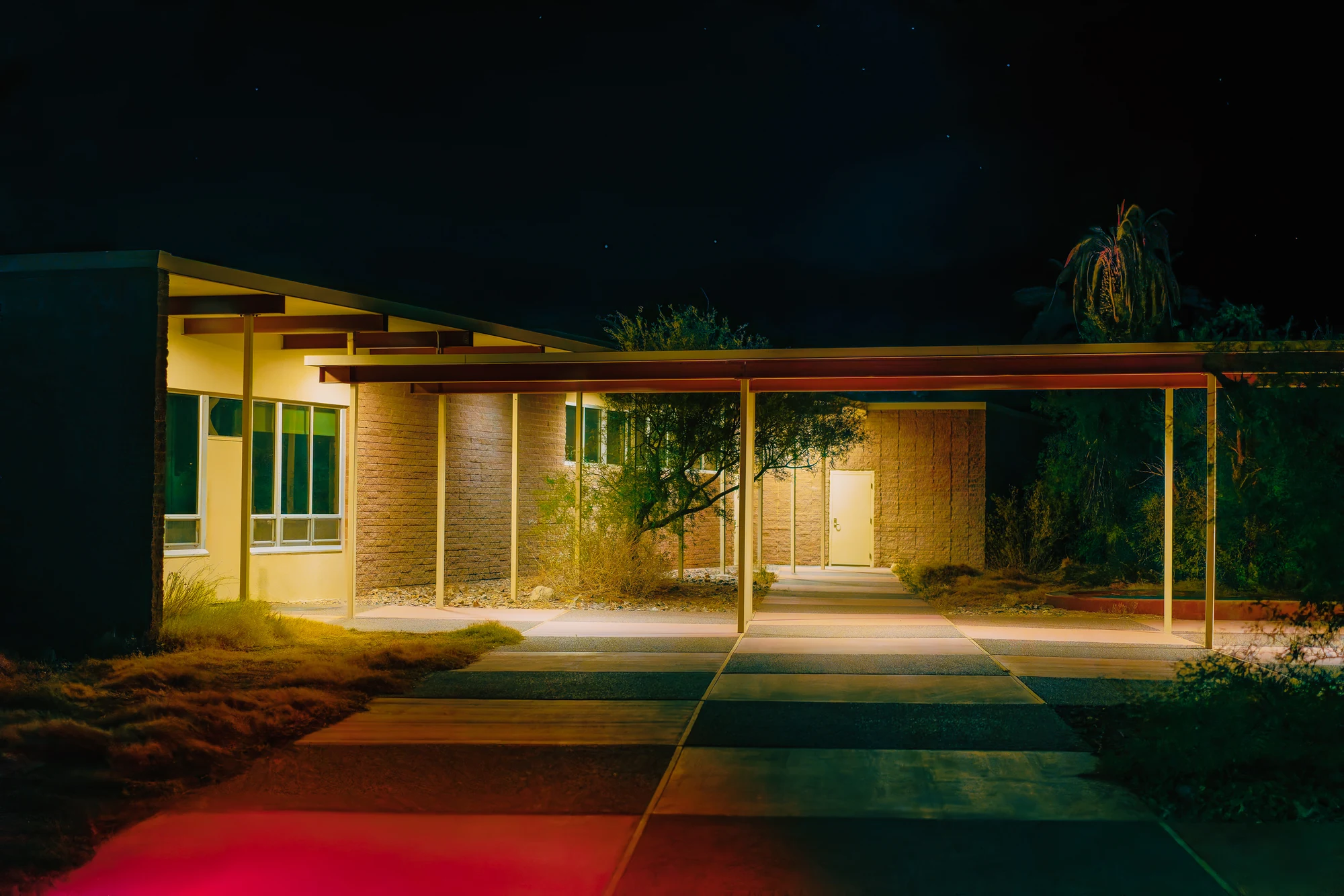 Low mid-century building with a long covered walkway lit at night, a red glow on the pavement in the foreground.