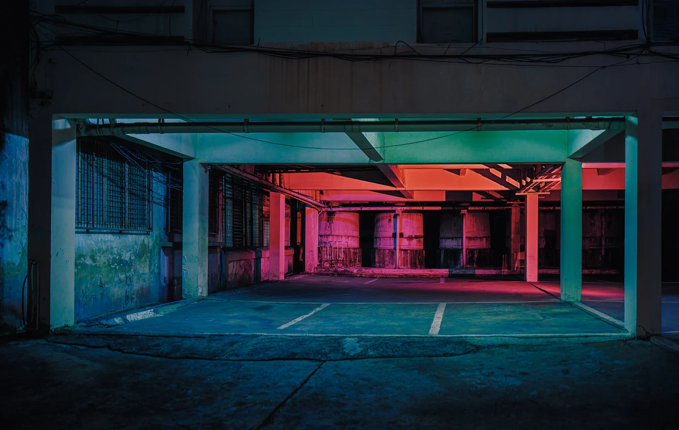 Parking vide au rez-de-chaussée d’un vieil immeuble, piliers éclairés en bleu canard devant et lumière rouge sur de grandes cuves au fond.
