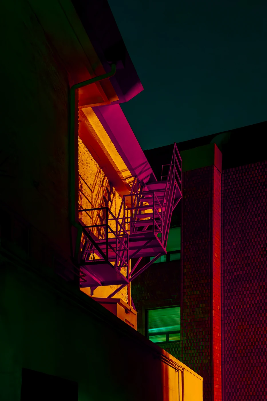 Escalier de secours métallique sur le flanc d’un immeuble éclairé en magenta contre un mur jaune, façade de brique rouge et ciel bleu canard.