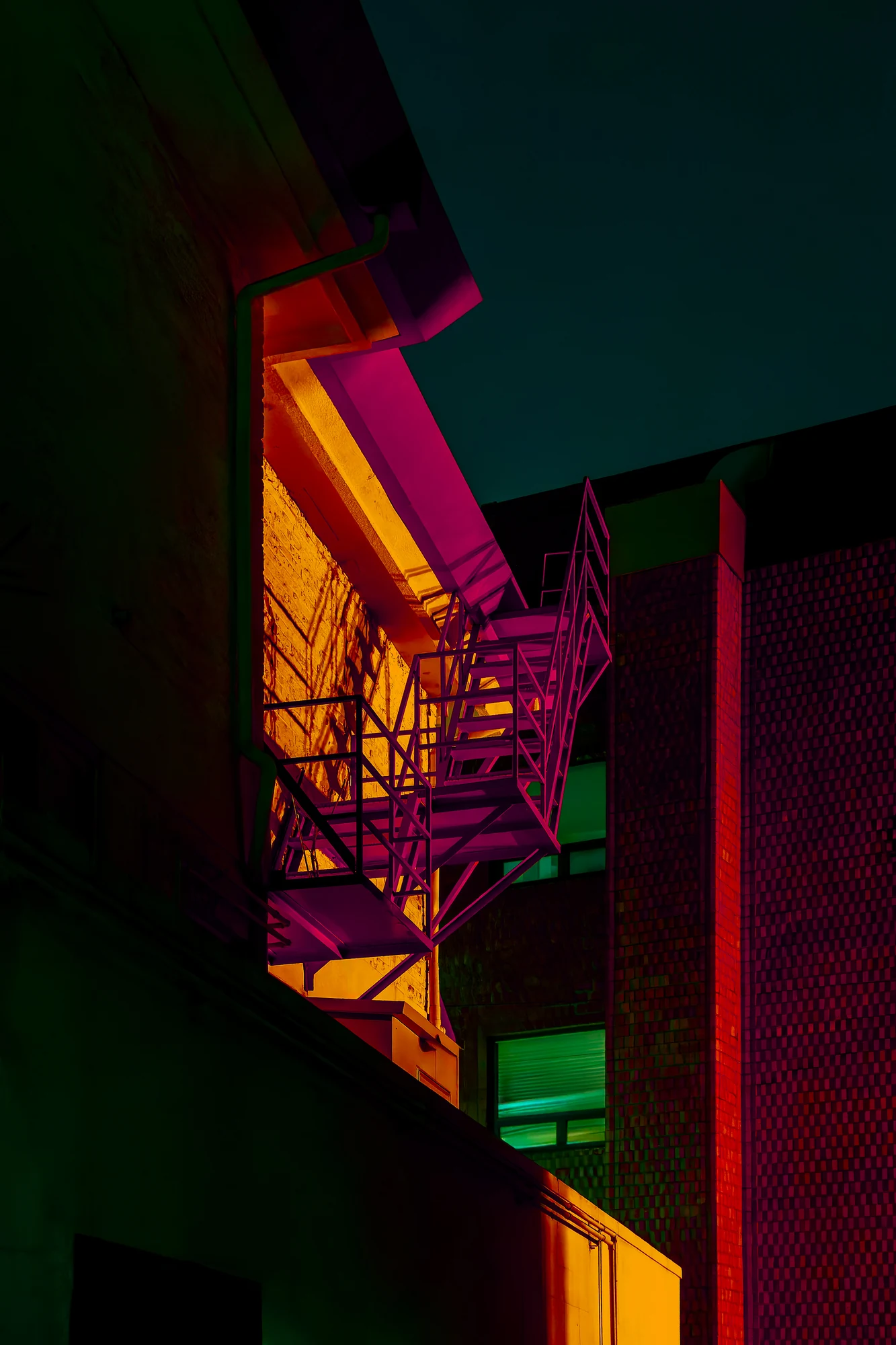 Metal fire escape on the side of a building lit magenta against a yellow wall, a red brick facade and teal sky behind.