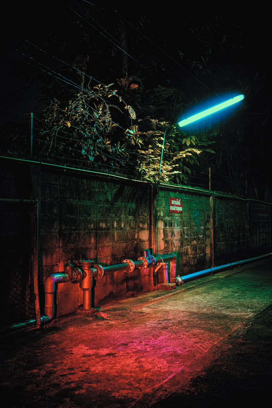 Pipes and valves along a stained block wall in a narrow alley at night, a cyan tube light above and a red glow on the ground.