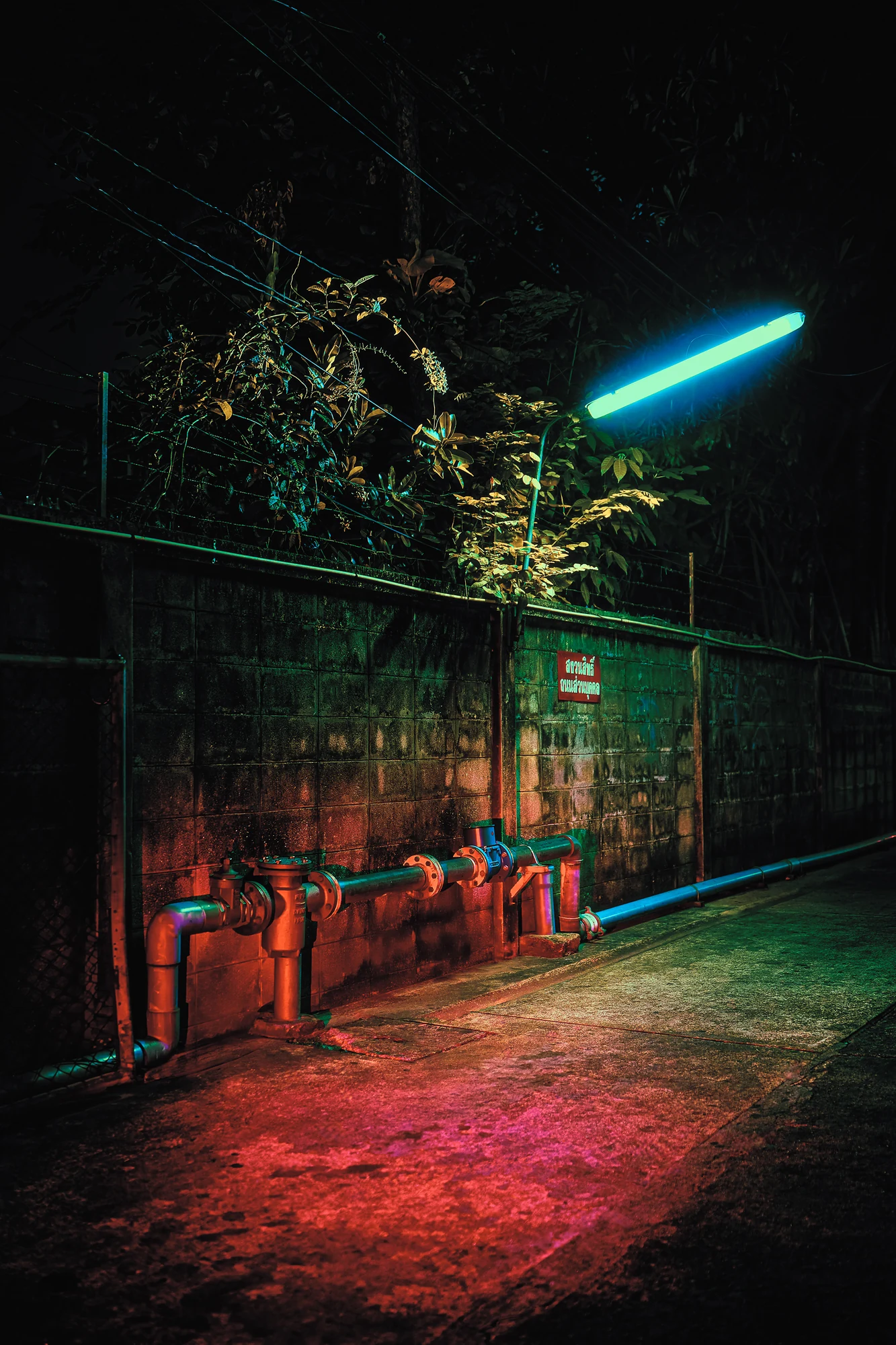 Pipes and valves along a stained block wall in a narrow alley at night, a cyan tube light above and a red glow on the ground.