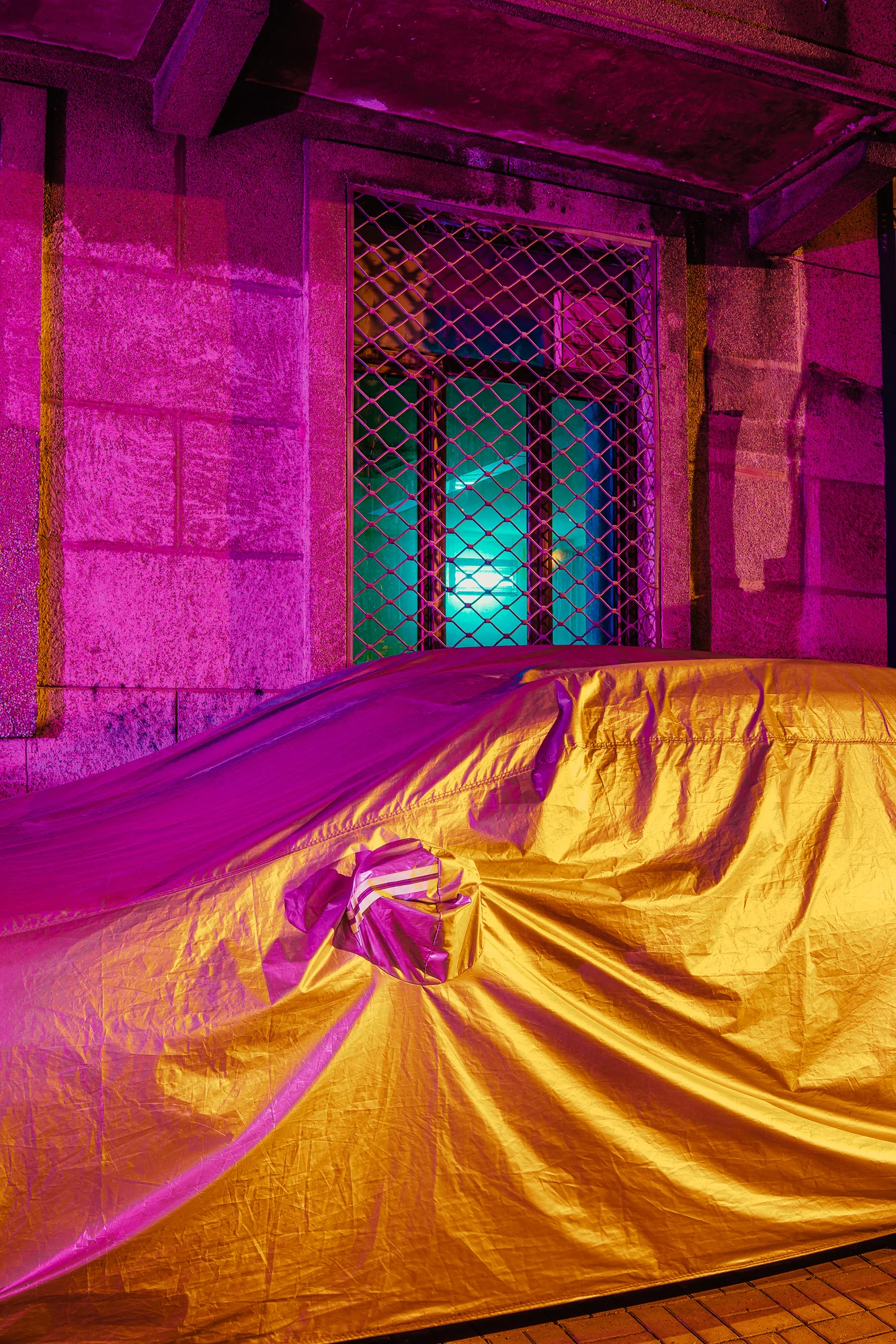 Car hidden under a shiny gold and magenta cover in front of a stone wall and a barred window glowing cyan.