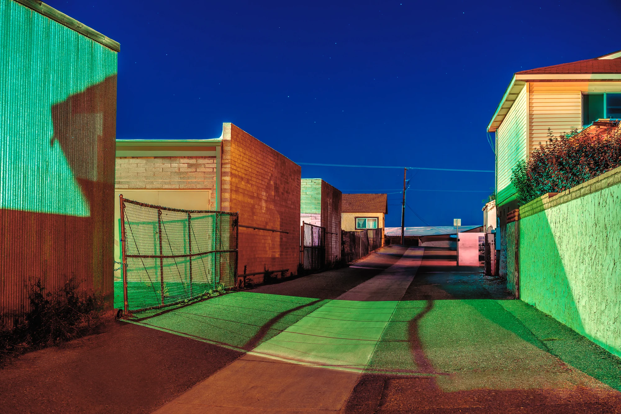 Ruelle vide la nuit éclairée en vert saturé et orange, un portail grillagé, des murs de brique et des maisons sous un ciel bleu étoilé.