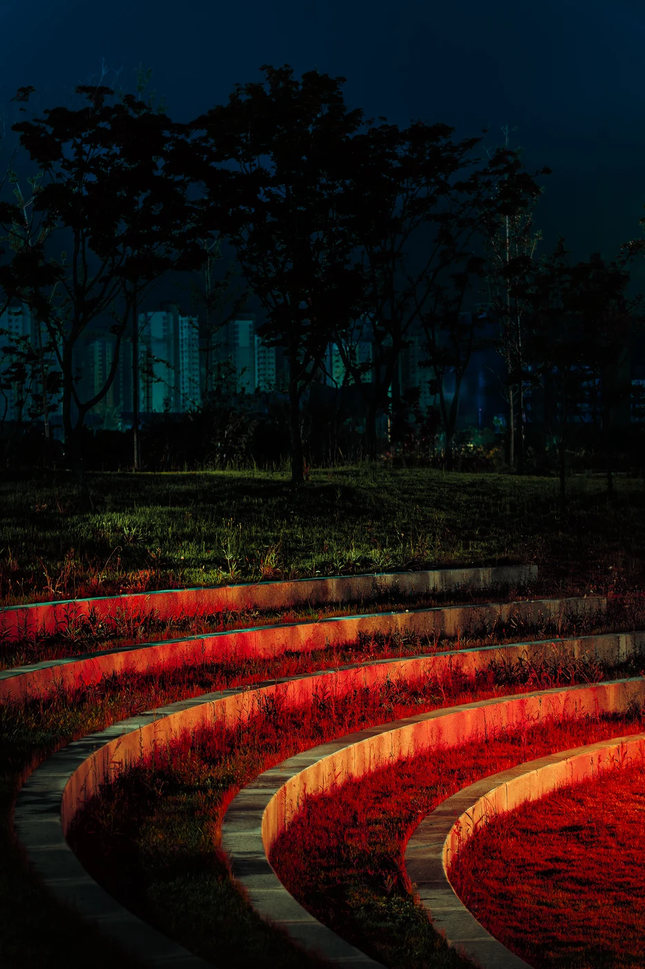Curved stone terraces glowing red in a park at night, dark trees and residential towers in a teal haze behind.