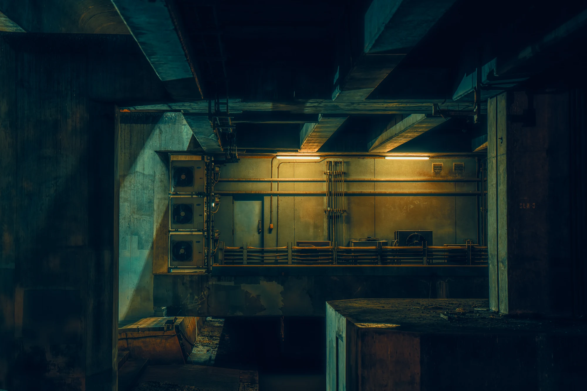Technical space under a concrete structure lit by two fluorescent tubes, with pipes, air-conditioning units and a grey door.