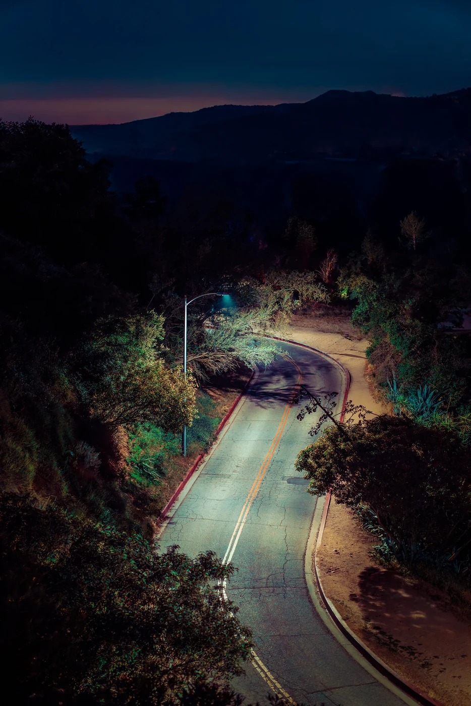 Route de colline sinueuse et vide vue d’en haut la nuit, éclairée par un seul lampadaire, collines sombres et lueur du crépuscule à l’horizon.