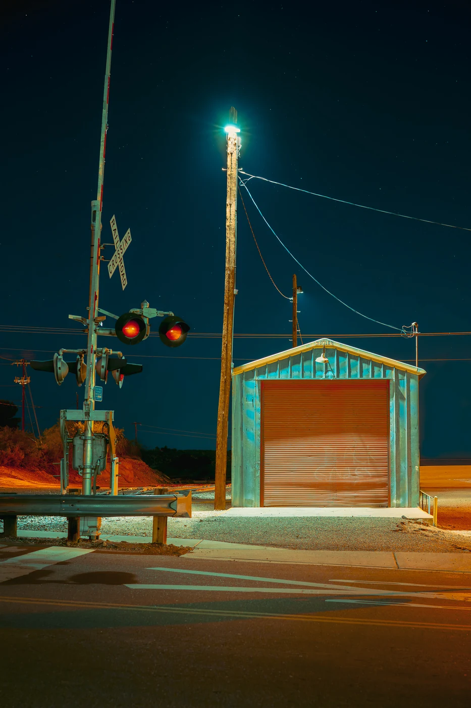 Railroad crossing signal with red lights beside a small metal shed with a roll-up door, lit by a single lamp on a wooden pole at night.