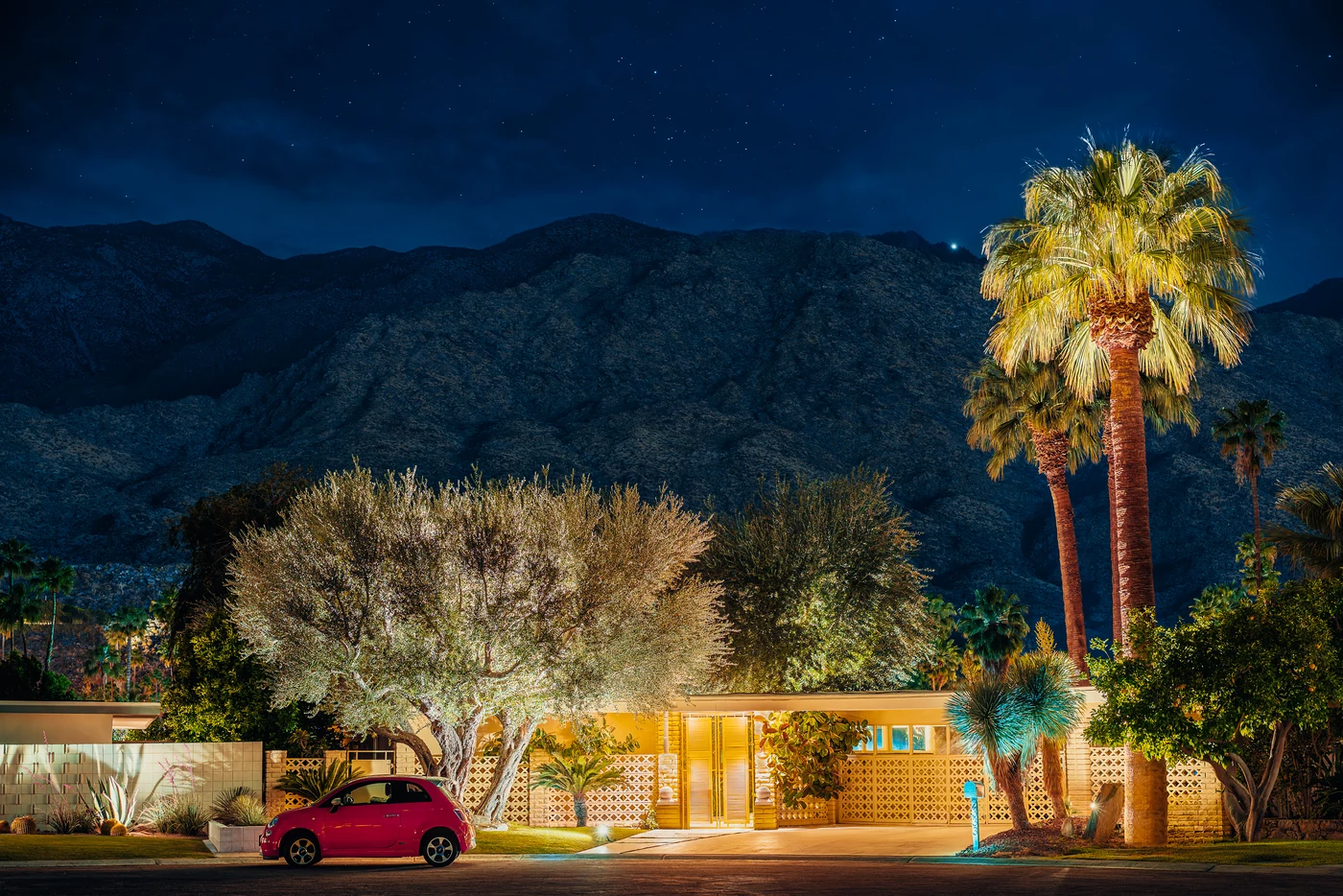 Mid-century modern house with breeze-block walls lit at night at the foot of dark mountains, palm and olive trees, a small pink car.
