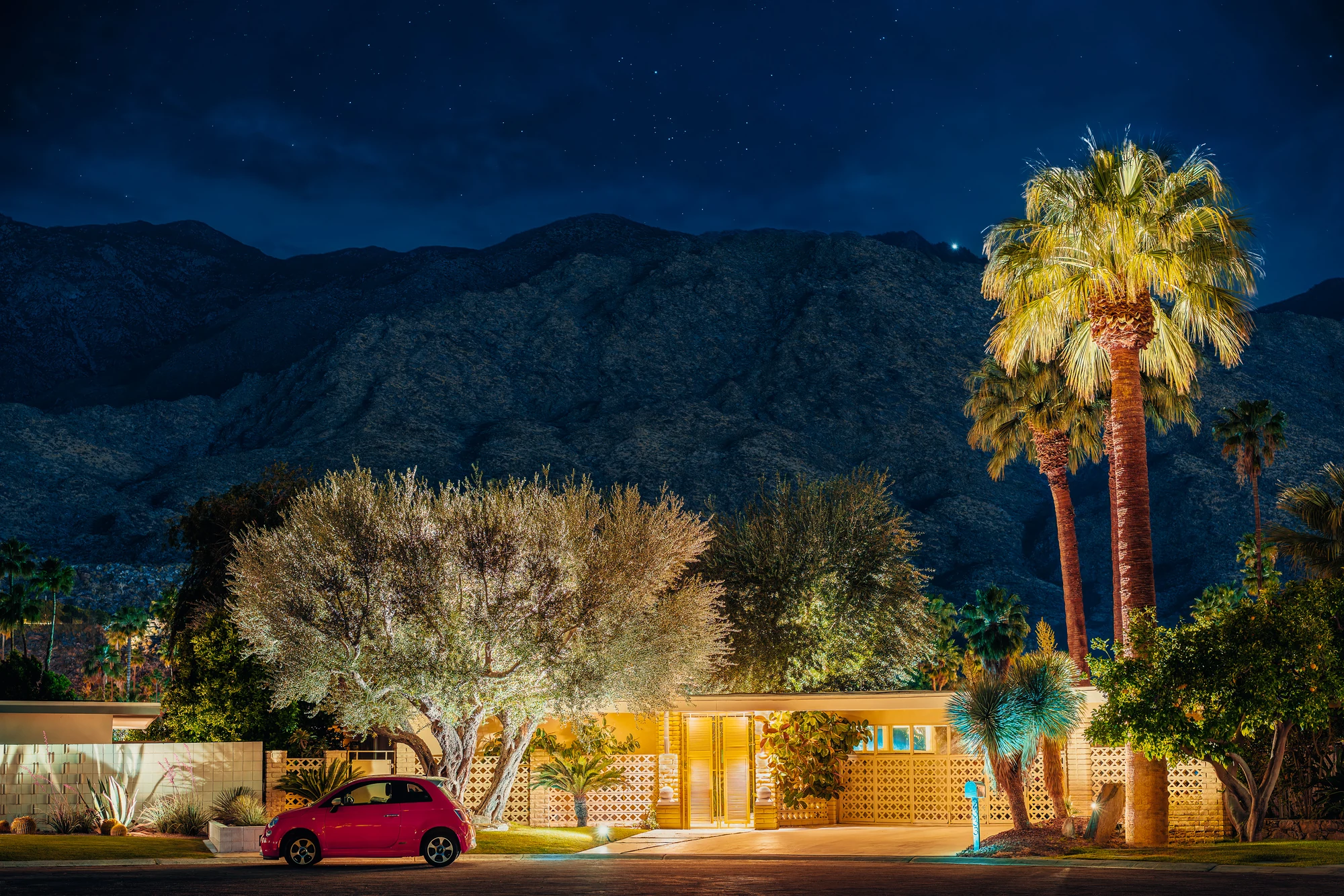 Mid-century modern house with breeze-block walls lit at night at the foot of dark mountains, palm and olive trees, a small pink car.