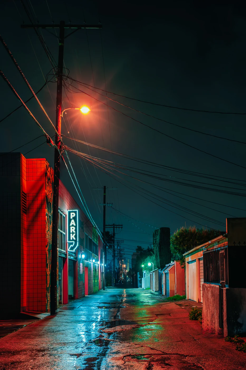 Ruelle mouillée la nuit avec une enseigne néon « PARK » sur un bâtiment rouge, un lampadaire orange et des lignes électriques enchevêtrées.