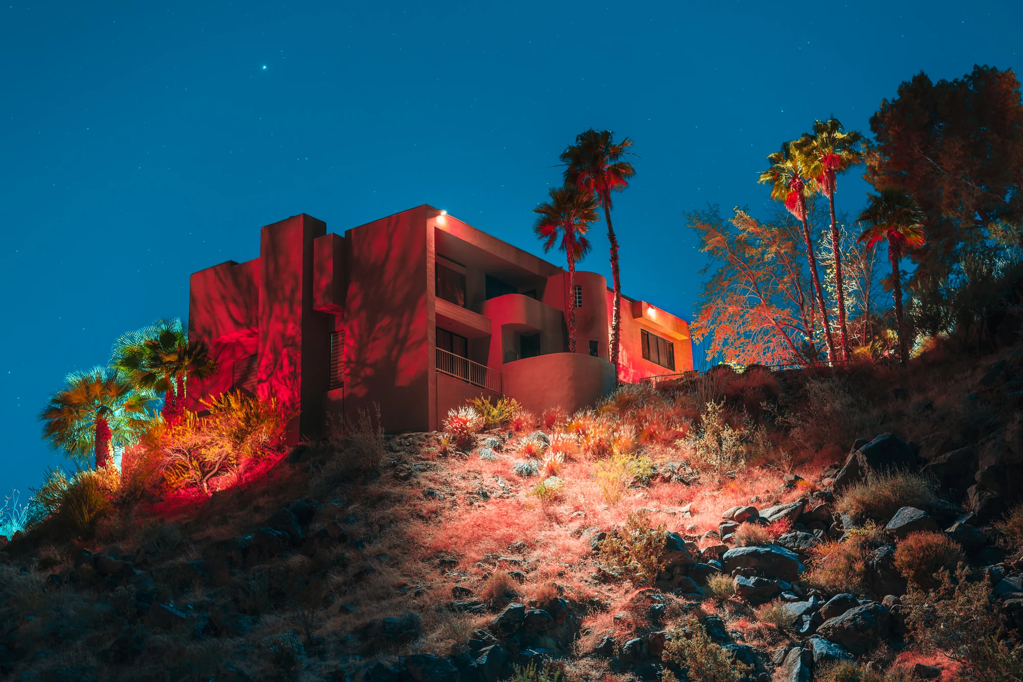 Geometric stucco house on a rocky hillside lit in red at night, palm trees and desert shrubs under a deep blue starry sky.