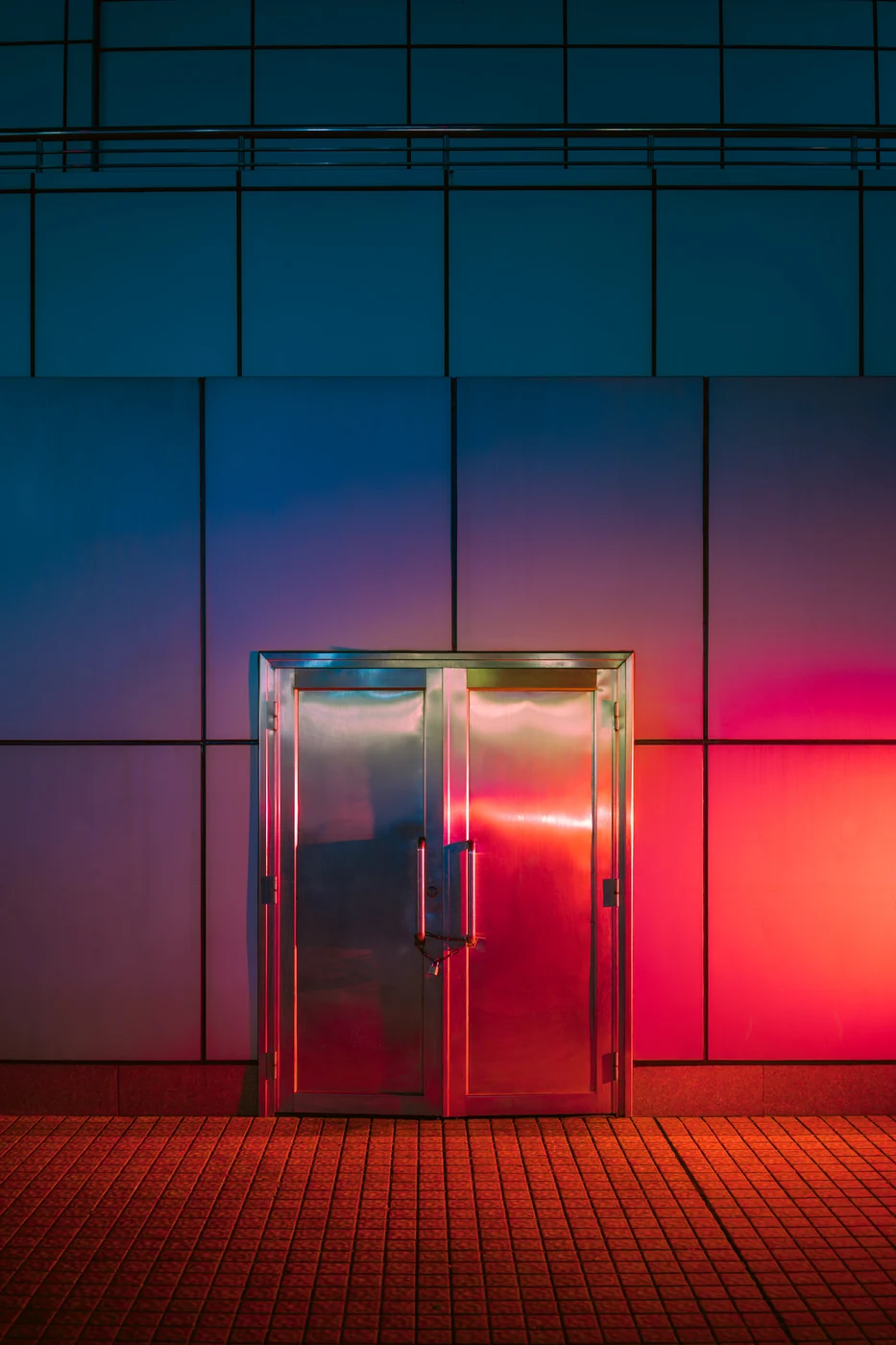 Double porte en acier poli dans une façade à panneaux éclairée d’un dégradé bleu, magenta et rouge, carrelage orange devant.