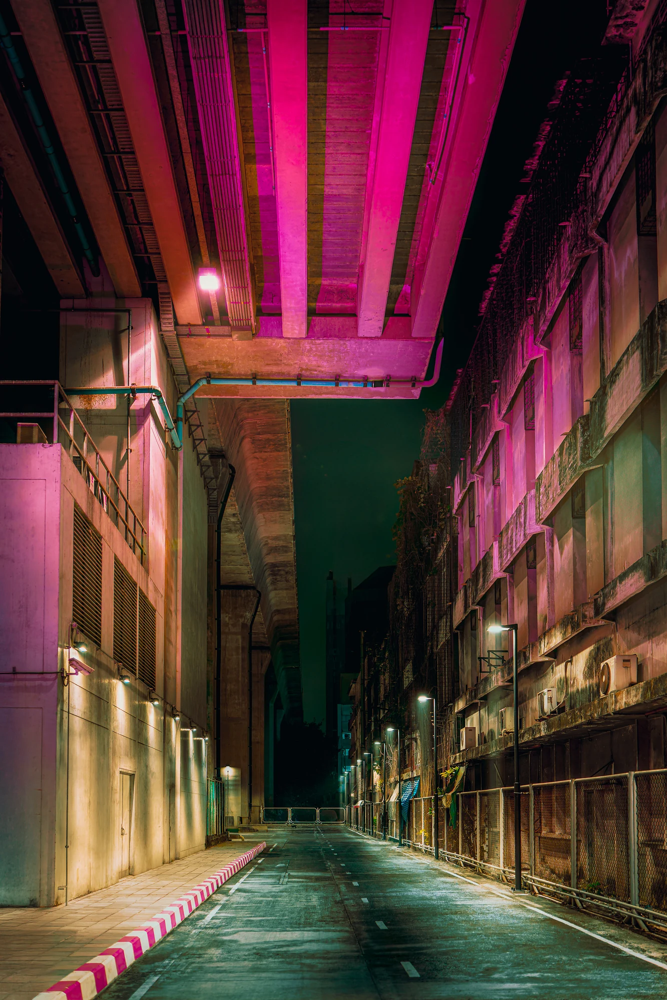 Empty wet street beneath an elevated structure glowing hot pink, an old grimy building with air-conditioners on the right.