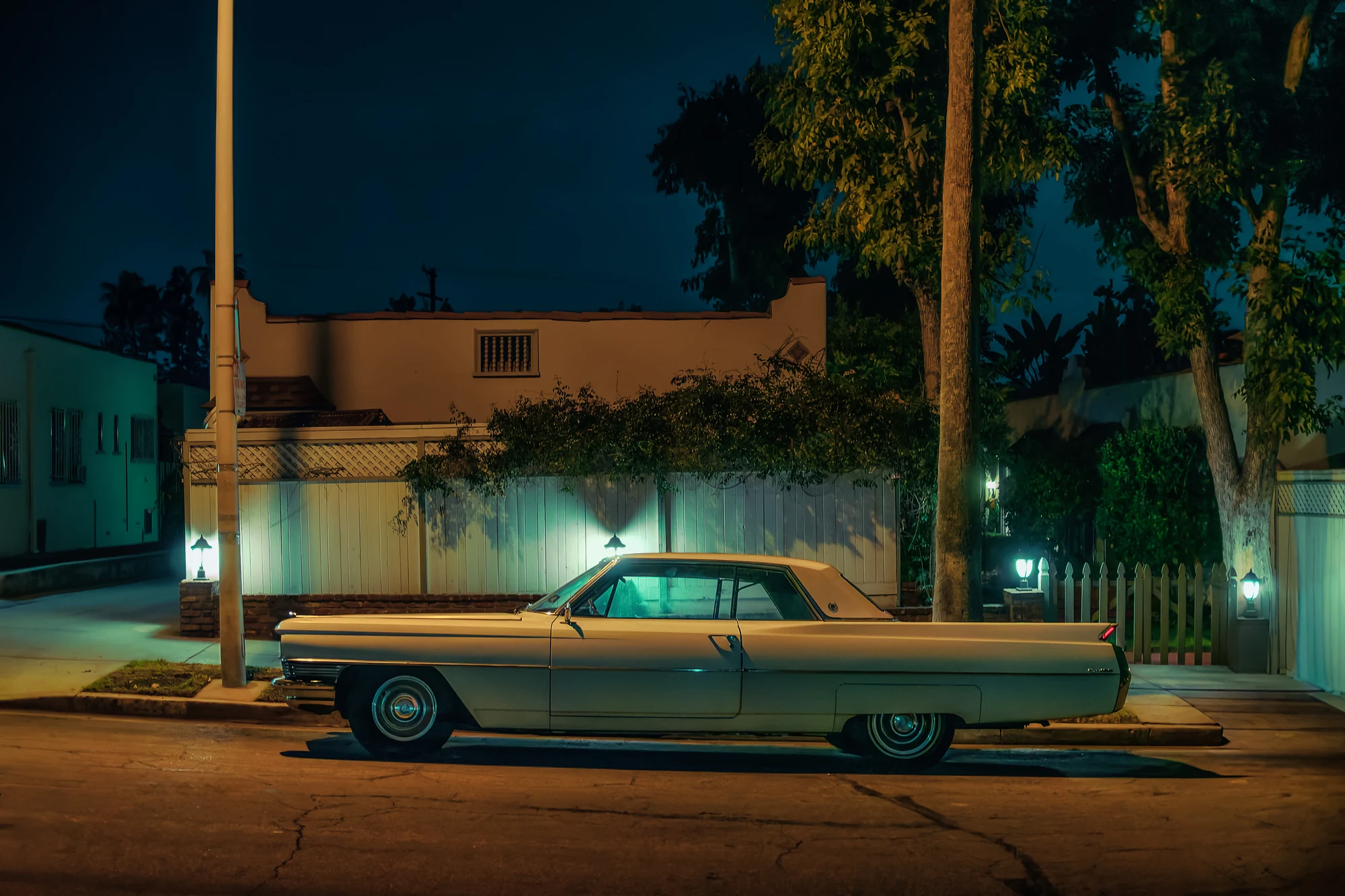 Pale vintage American coupé with tail fins parked in profile on a dark residential street, garden lamps and a white fence behind.