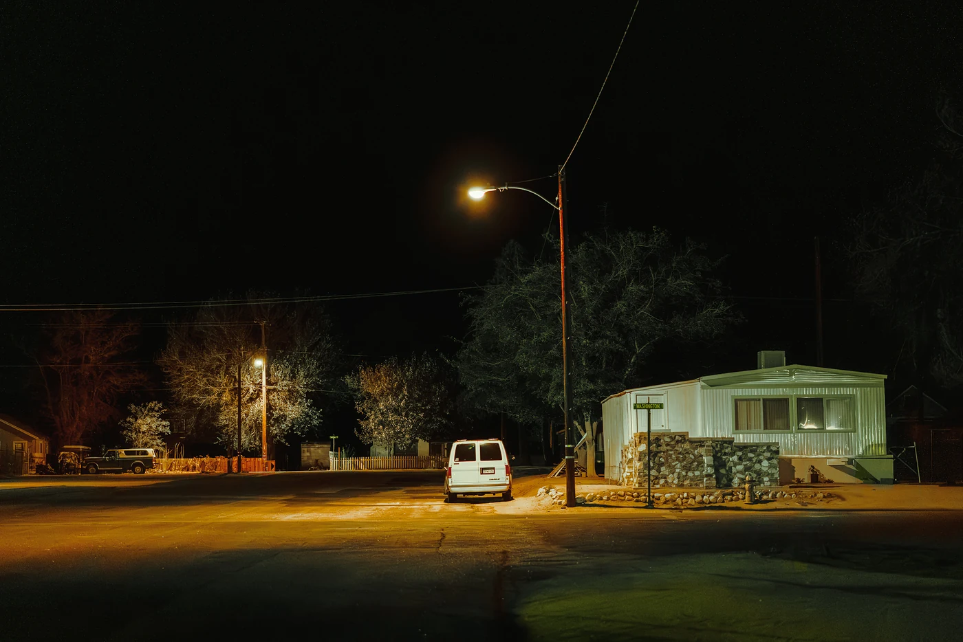 White van parked under a streetlight beside a white mobile home at night, bare trees and a quiet residential street behind.