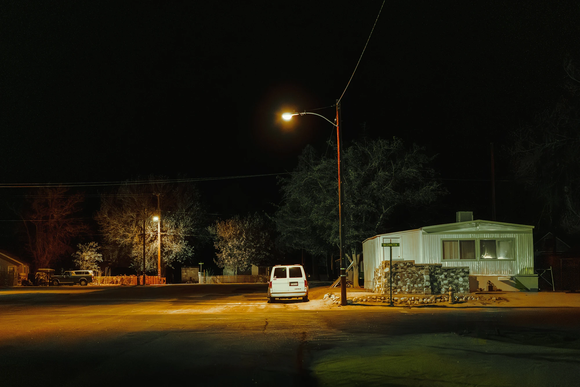 White van parked under a streetlight beside a white mobile home at night, bare trees and a quiet residential street behind.