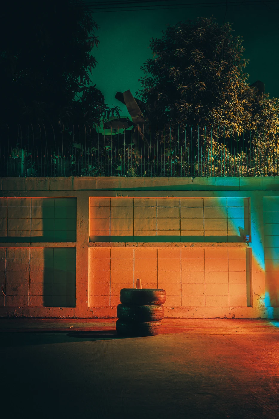 Stack of three old tyres against a tiled wall lit orange and cyan at night, railings and trees above.