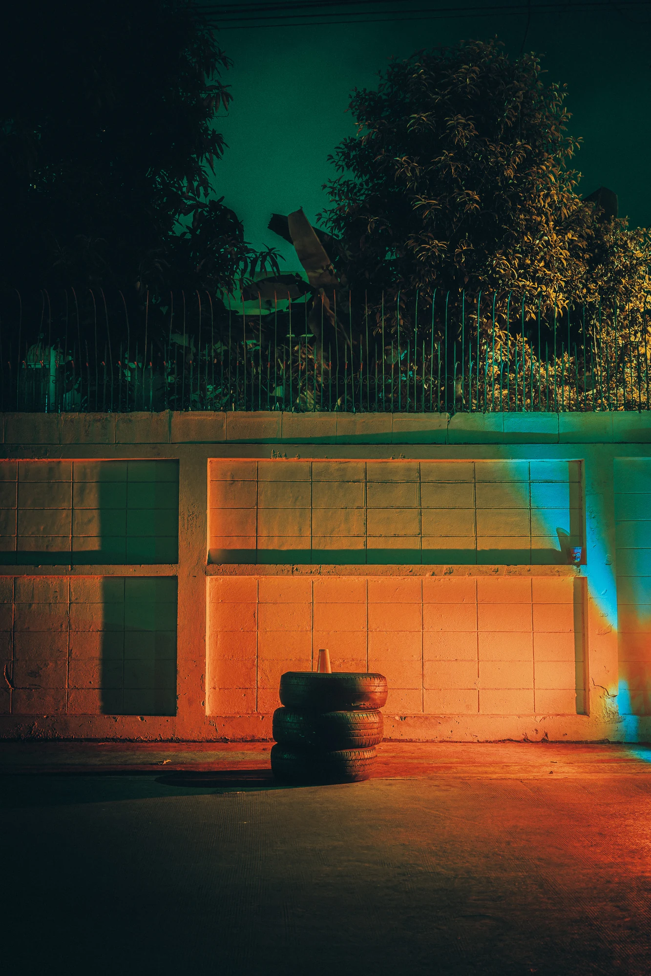 Stack of three old tyres against a tiled wall lit orange and cyan at night, railings and trees above.