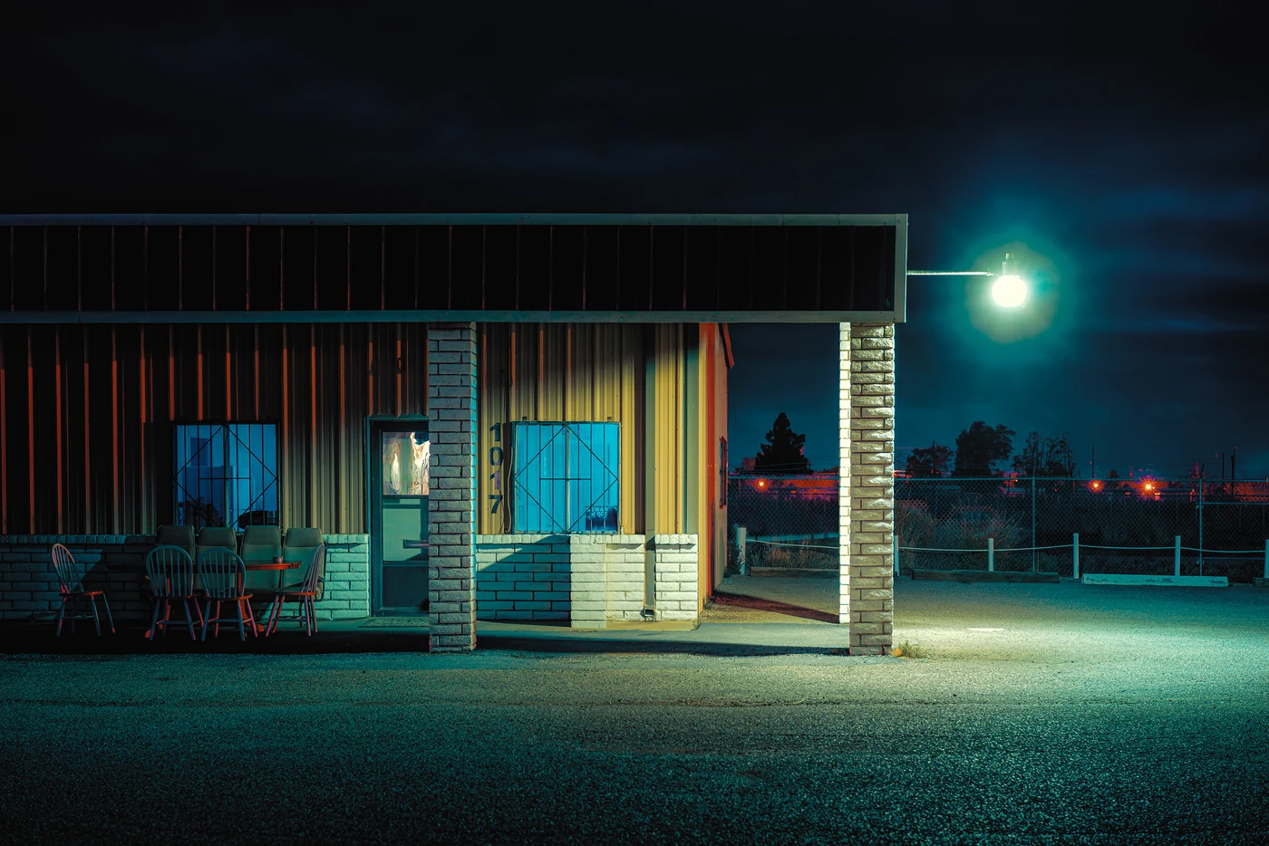 Porch of a low metal building at night with red chairs around a table, a glowing lamp lighting an empty gravel lot.