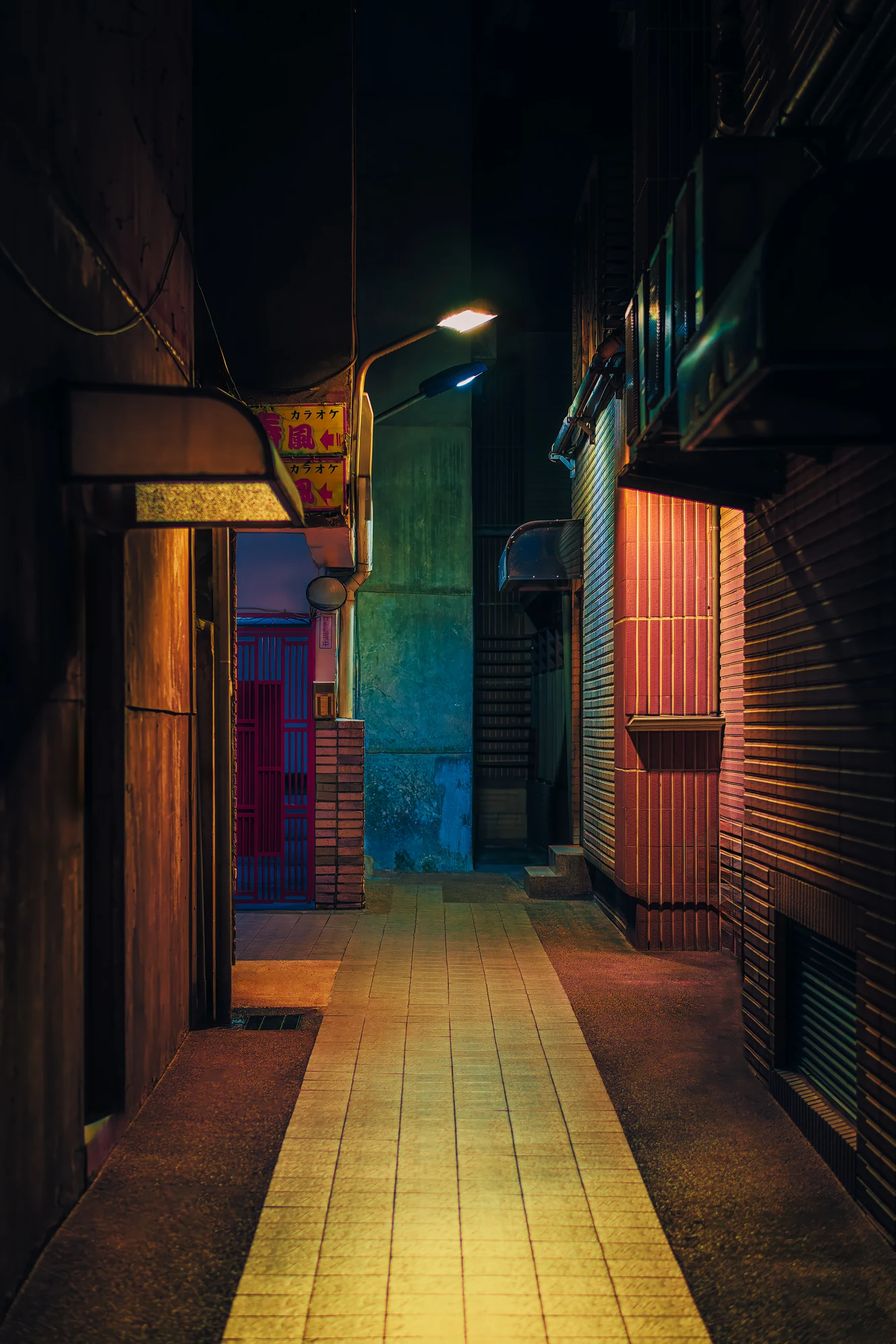 Narrow tiled lane between buildings at night leading to a karaoke sign and a red-grilled door, a streetlight overhead.