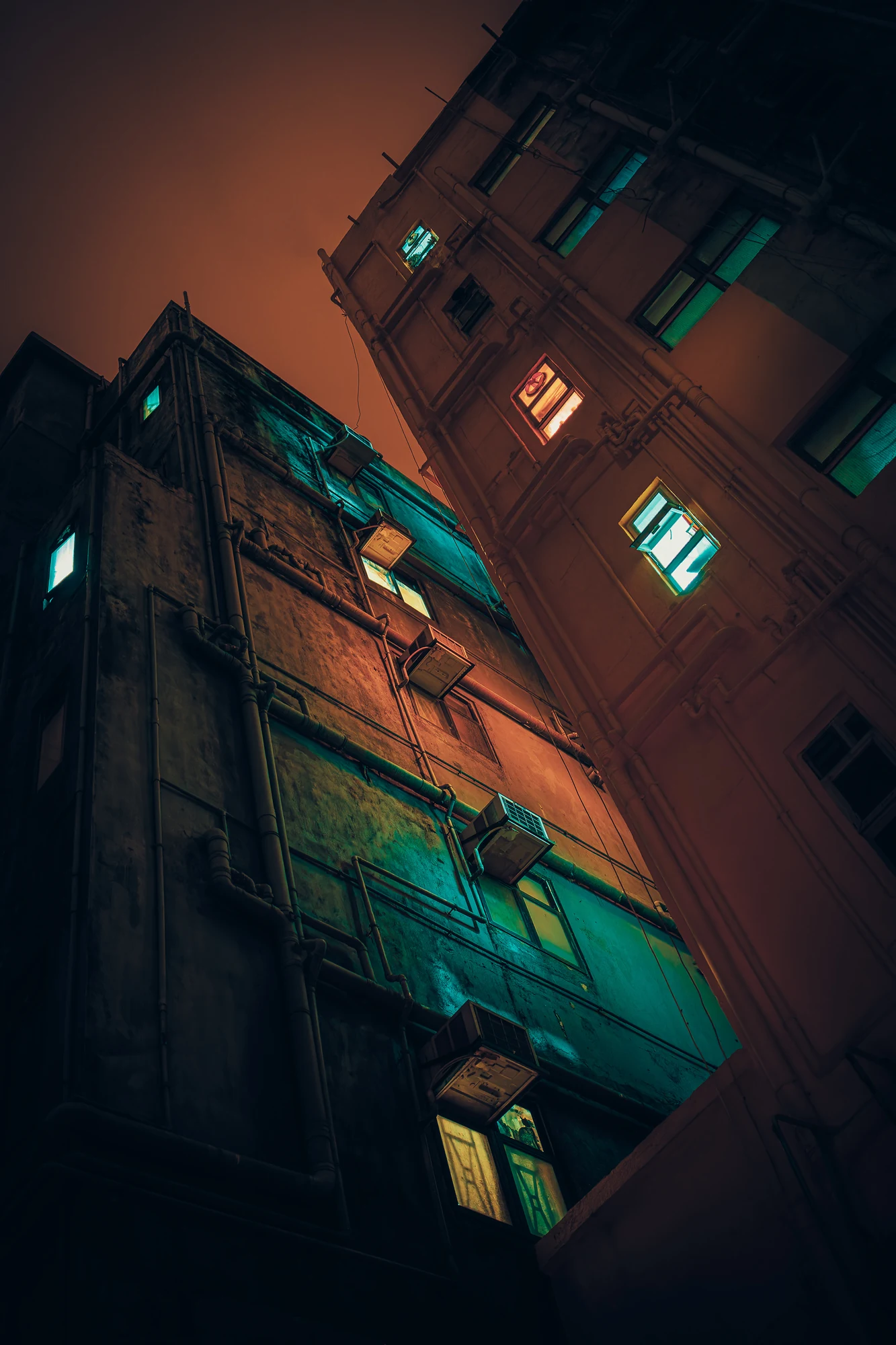 Upward view of two old apartment blocks with air-conditioners and a few lit windows in green and orange under a reddish sky.