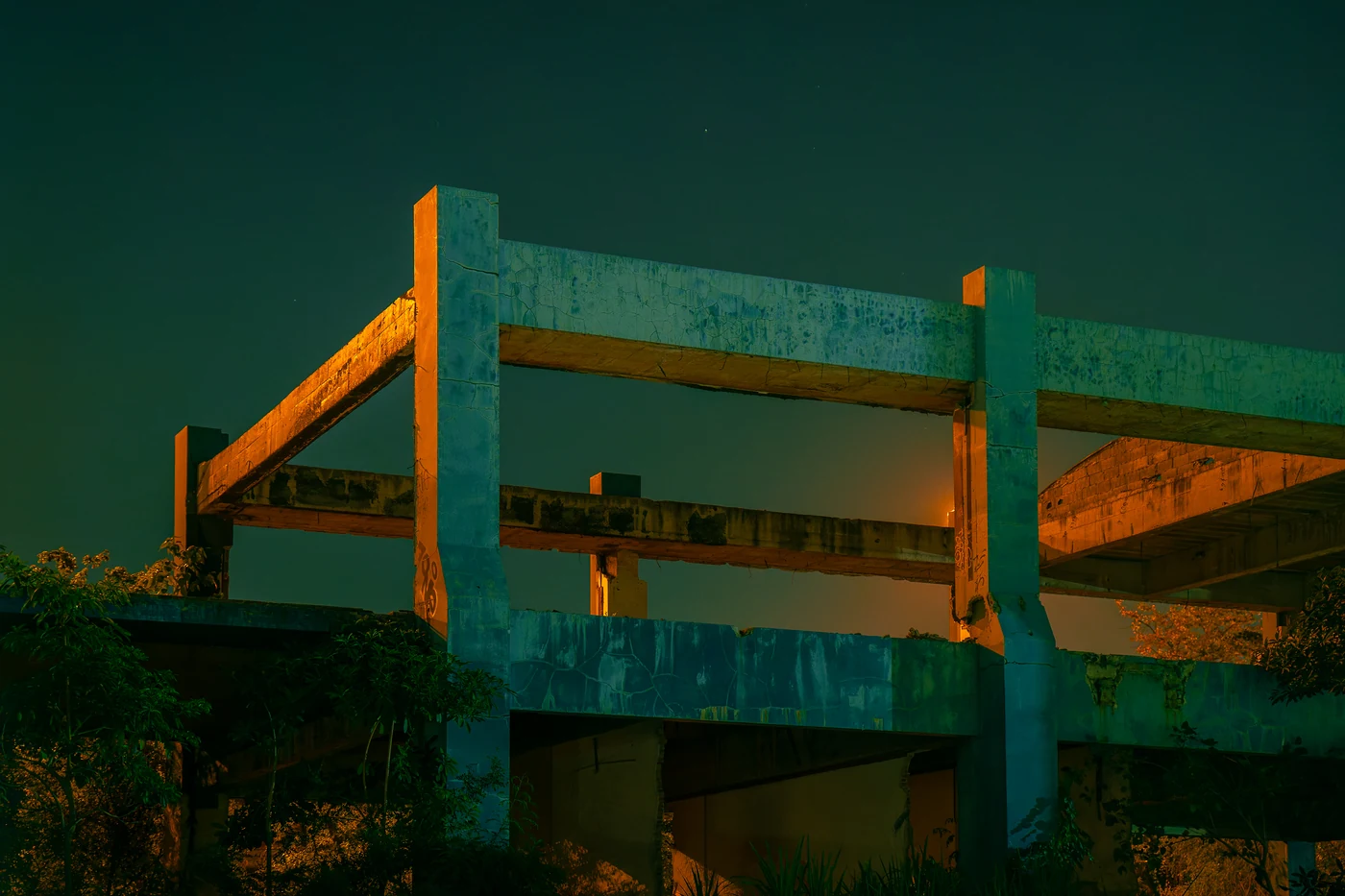 Bare concrete frame of an abandoned unfinished building lit teal and orange at night above trees.
