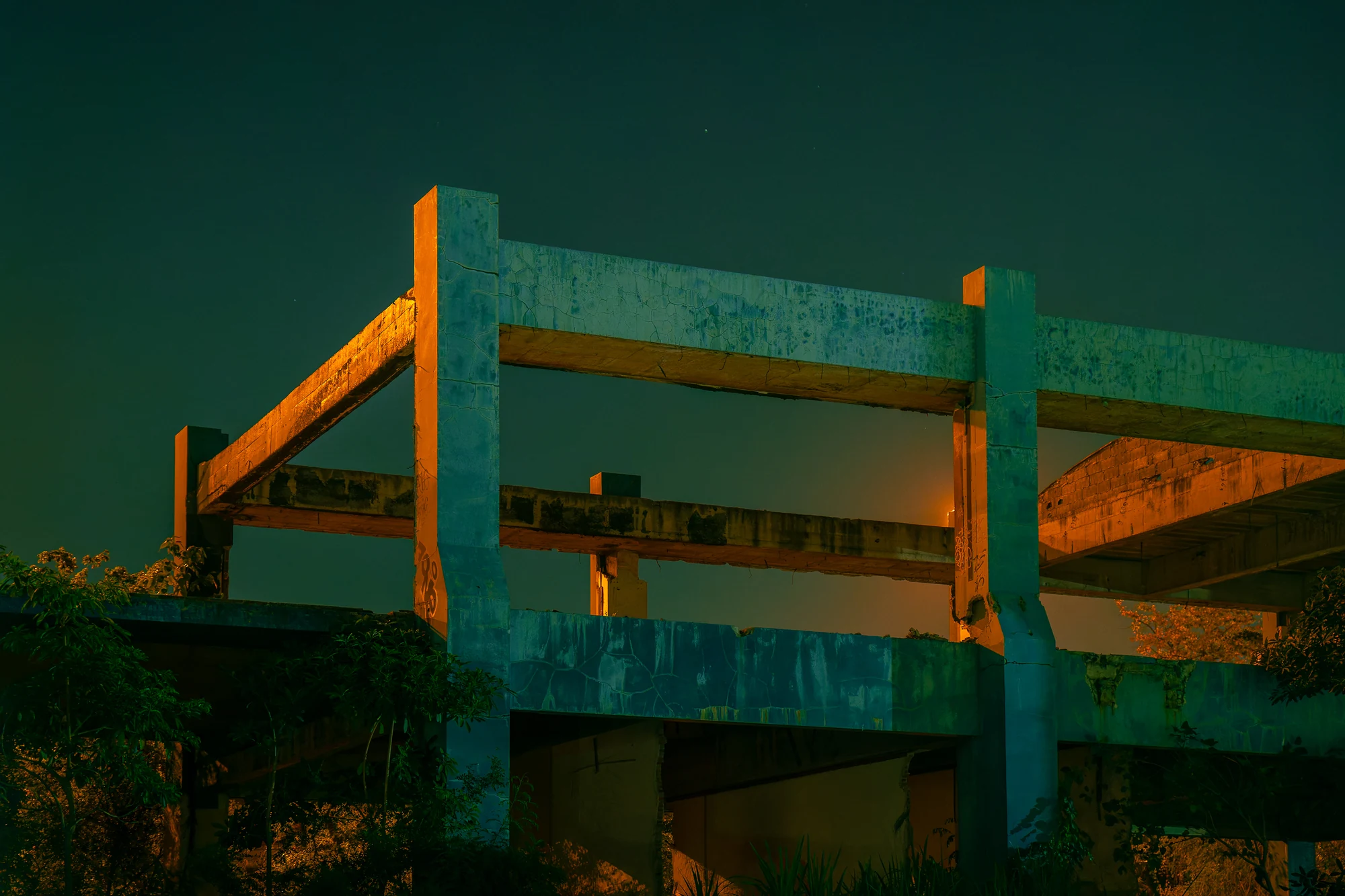 Bare concrete frame of an abandoned unfinished building lit teal and orange at night above trees.