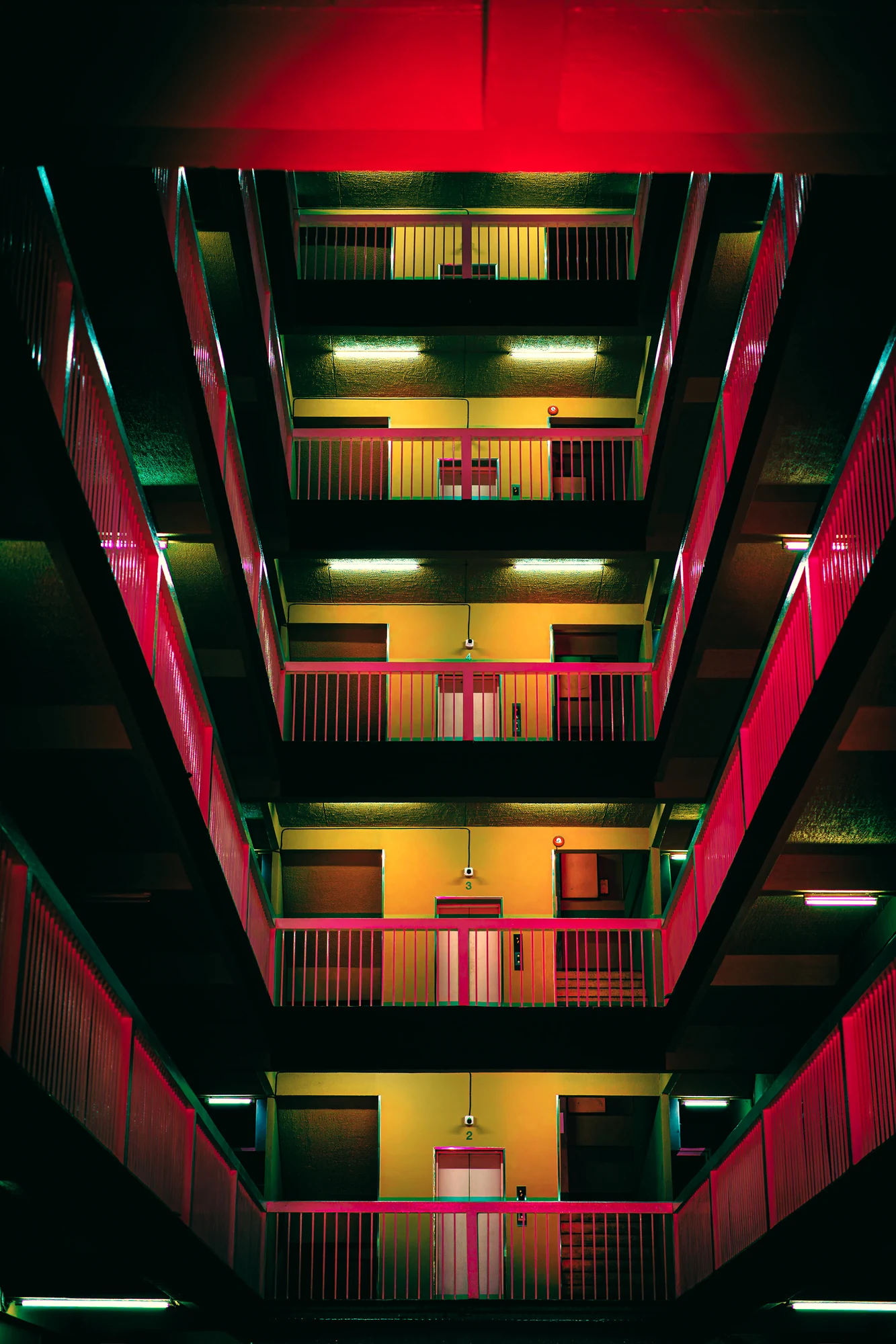 Vue symétrique vers le haut d’un atrium d’immeuble, garde-corps roses, murs jaunes et portes répétés à chaque étage, plafond rouge.