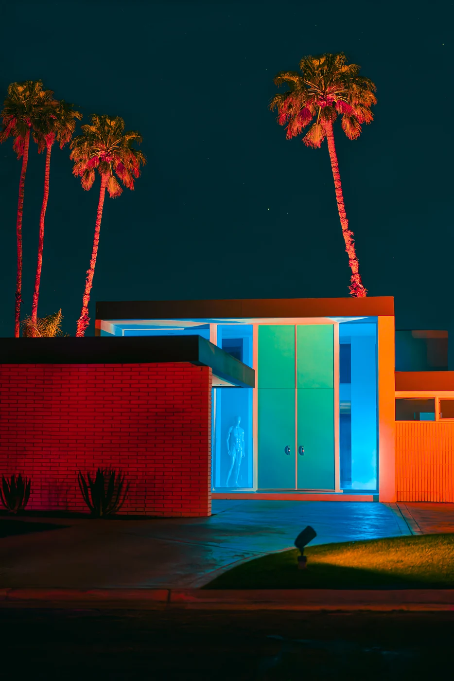 Entrée de maison moderniste la nuit, double porte verte, panneaux de verre bleu lumineux avec une silhouette pâle derrière, mur de brique rouge et palmiers.