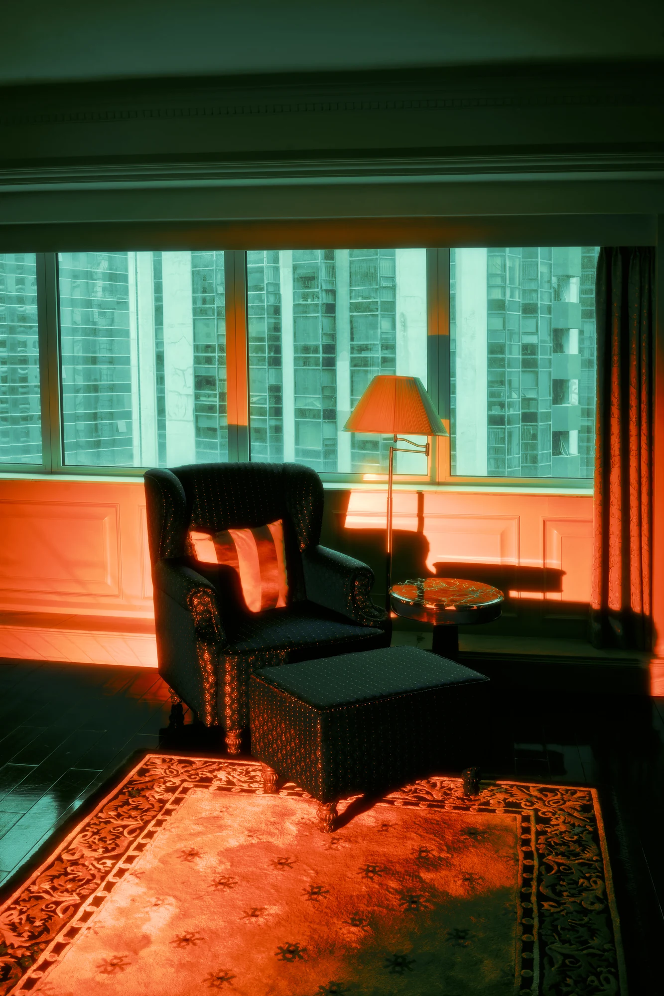 Hotel room corner with an armchair, footstool and standing lamp in warm orange light, a wall of towers seen through the window.