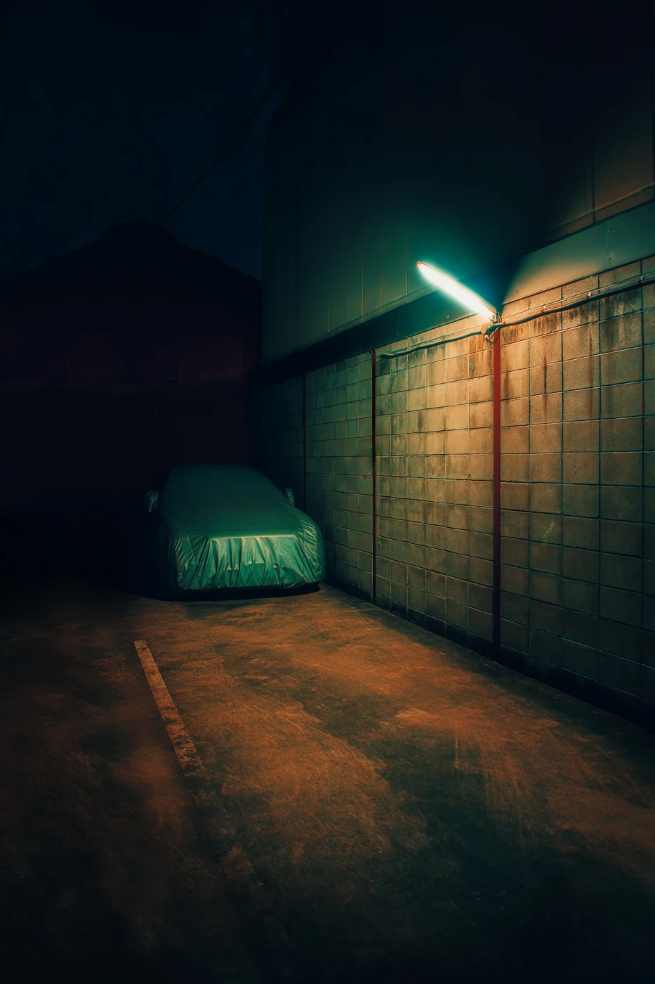 Voiture sous une housse de protection sur une place de parking sombre, contre un mur carrelé éclairé par un unique néon.